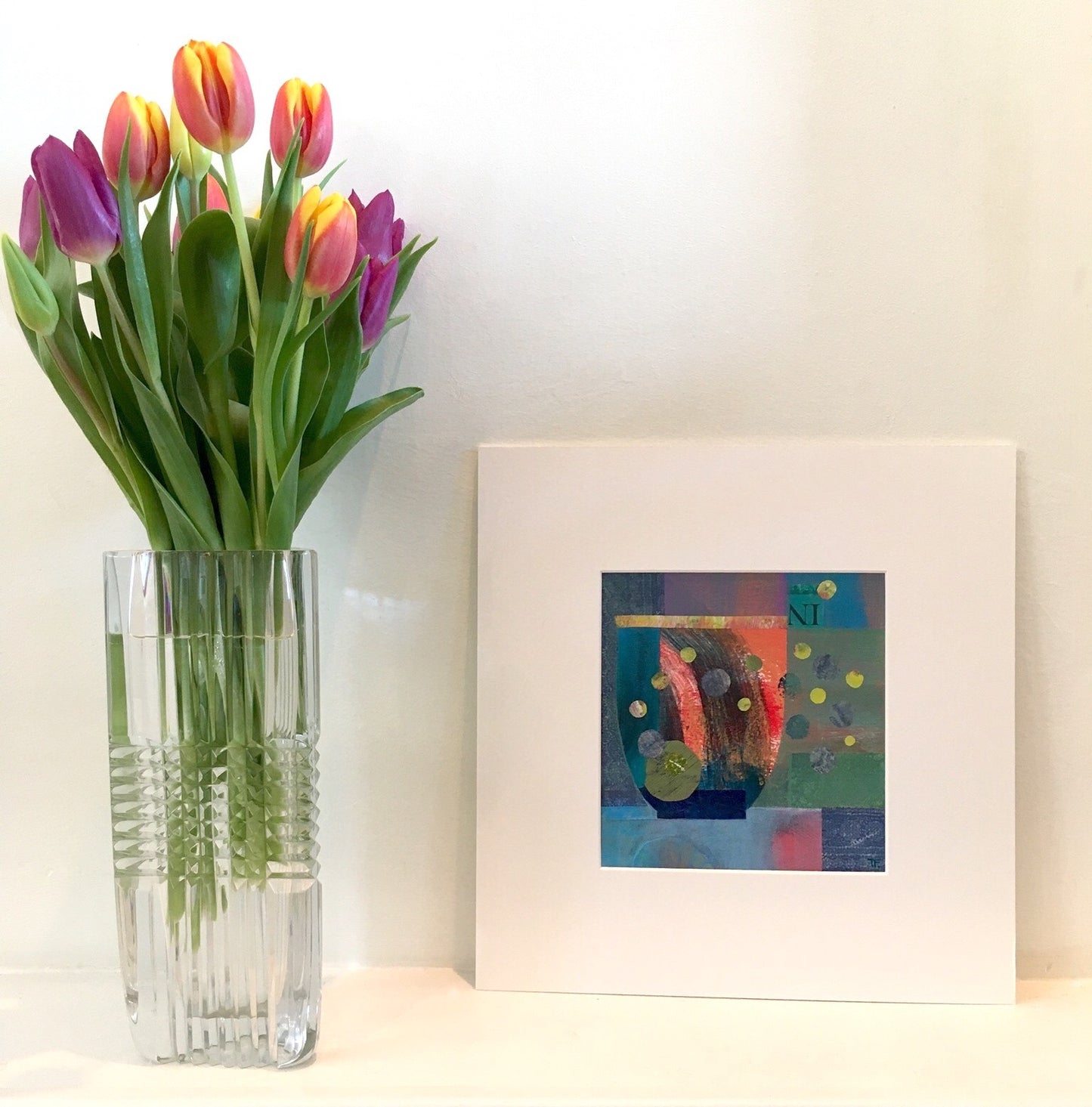 A Wave Inside by Teresa Flavin Artist, abstract still life mixed media painting shown in white mount on shelf next to tulips in vase.