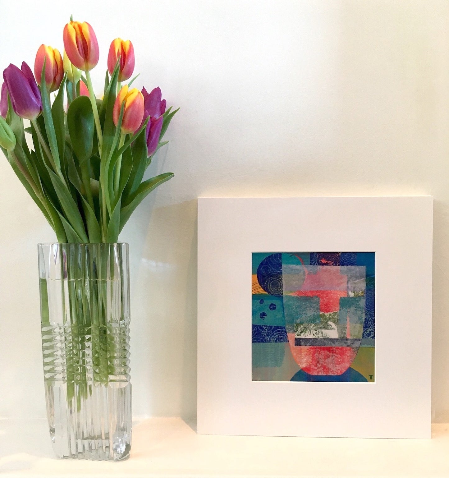 The Warmth Inside by Teresa Flavin Artist, abstract still life mixed media painting shown in white mount on shelf next to tulips in vase.