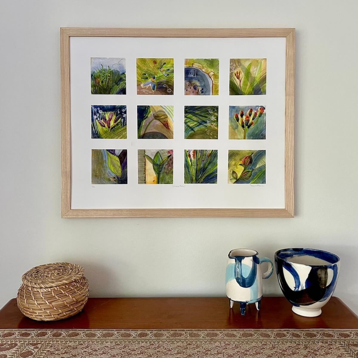 Summer Rain, abstract botanical art print by artist Teresa Flavin, flower forms, leaves and stems in gold, greens, orange, blue and grey, shown in light wood frame on wall.