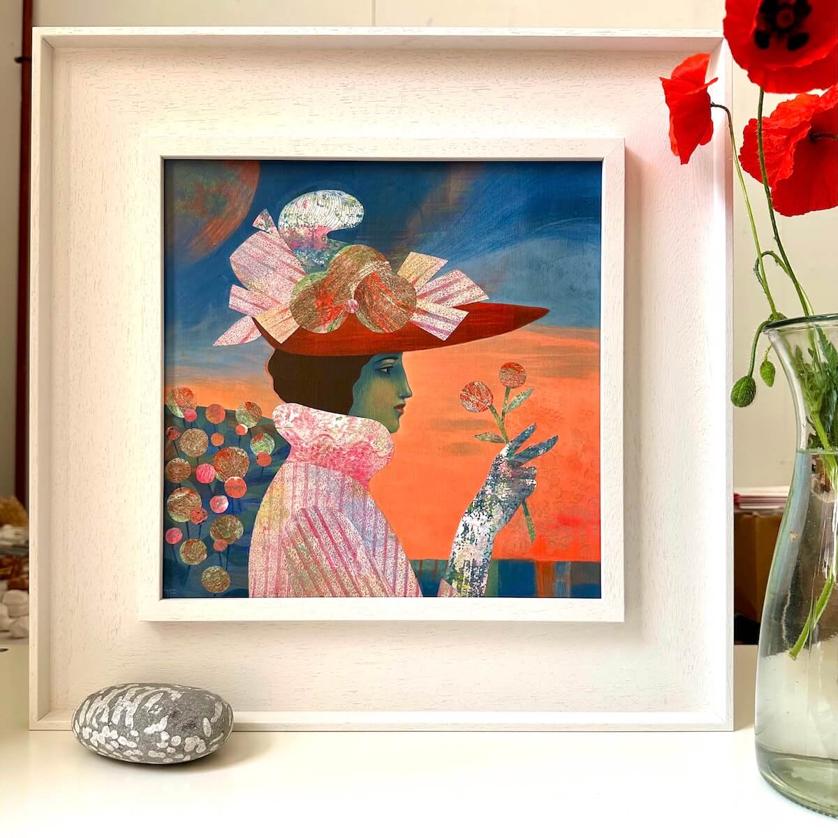 White framed Aurore painting by Teresa Flavin on a table next to  a stone and poppies in a vase.