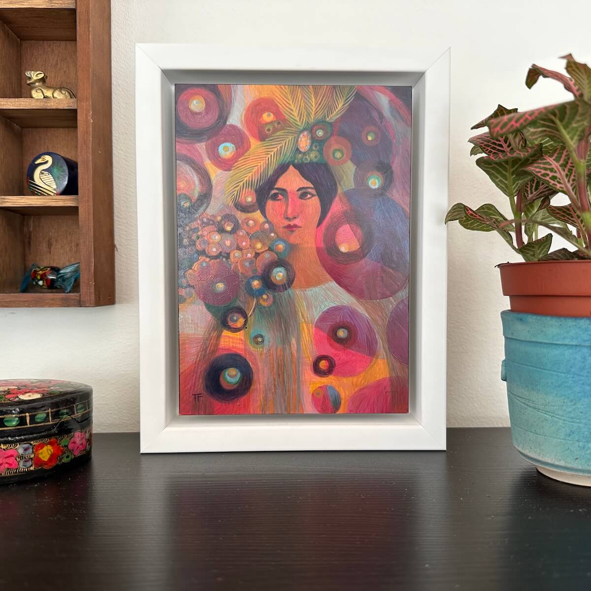 Colorful abstract art print in a white frame on a dark surface with a plant and shelves in the background.