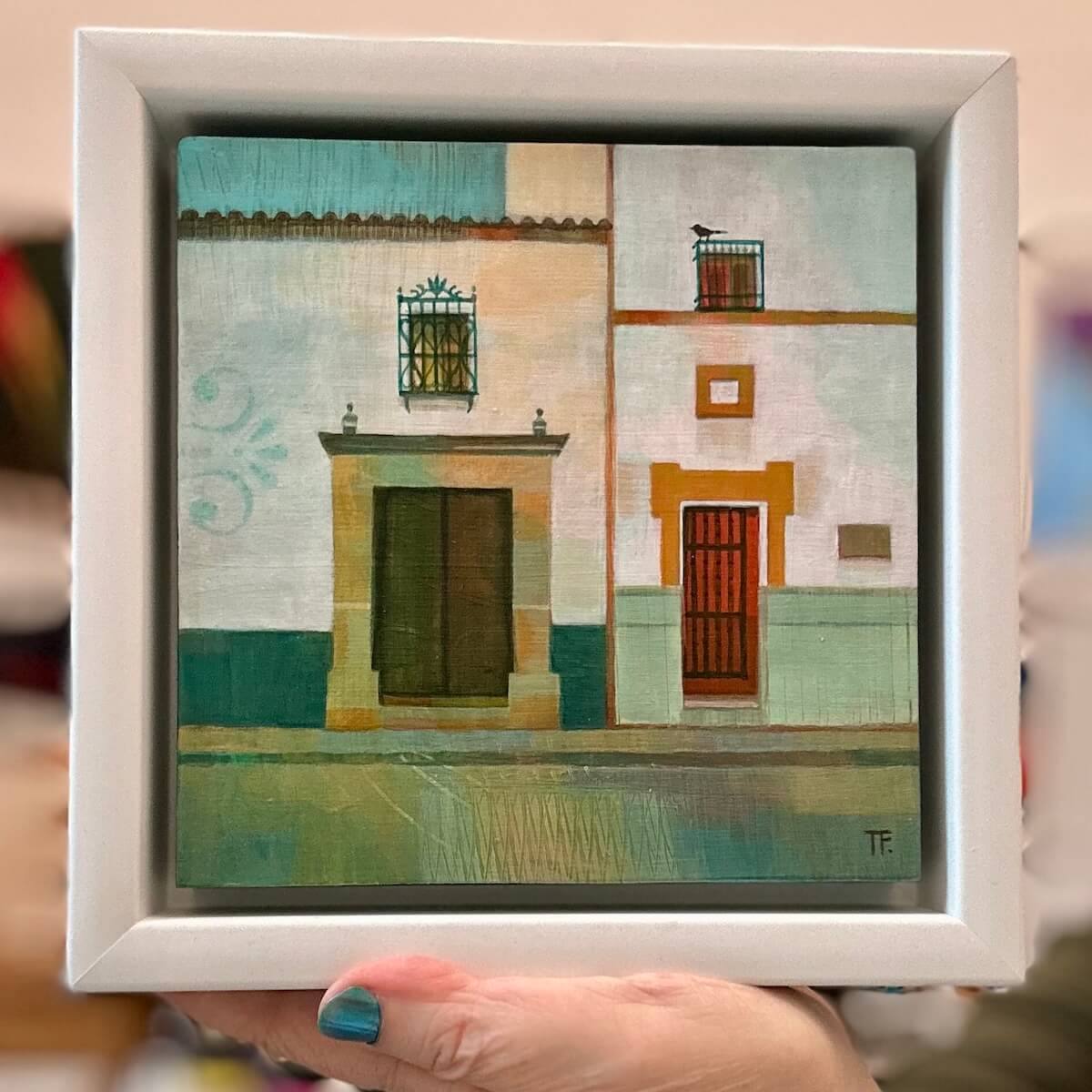 Small painting of a Spanish street scene  held in a white frame.