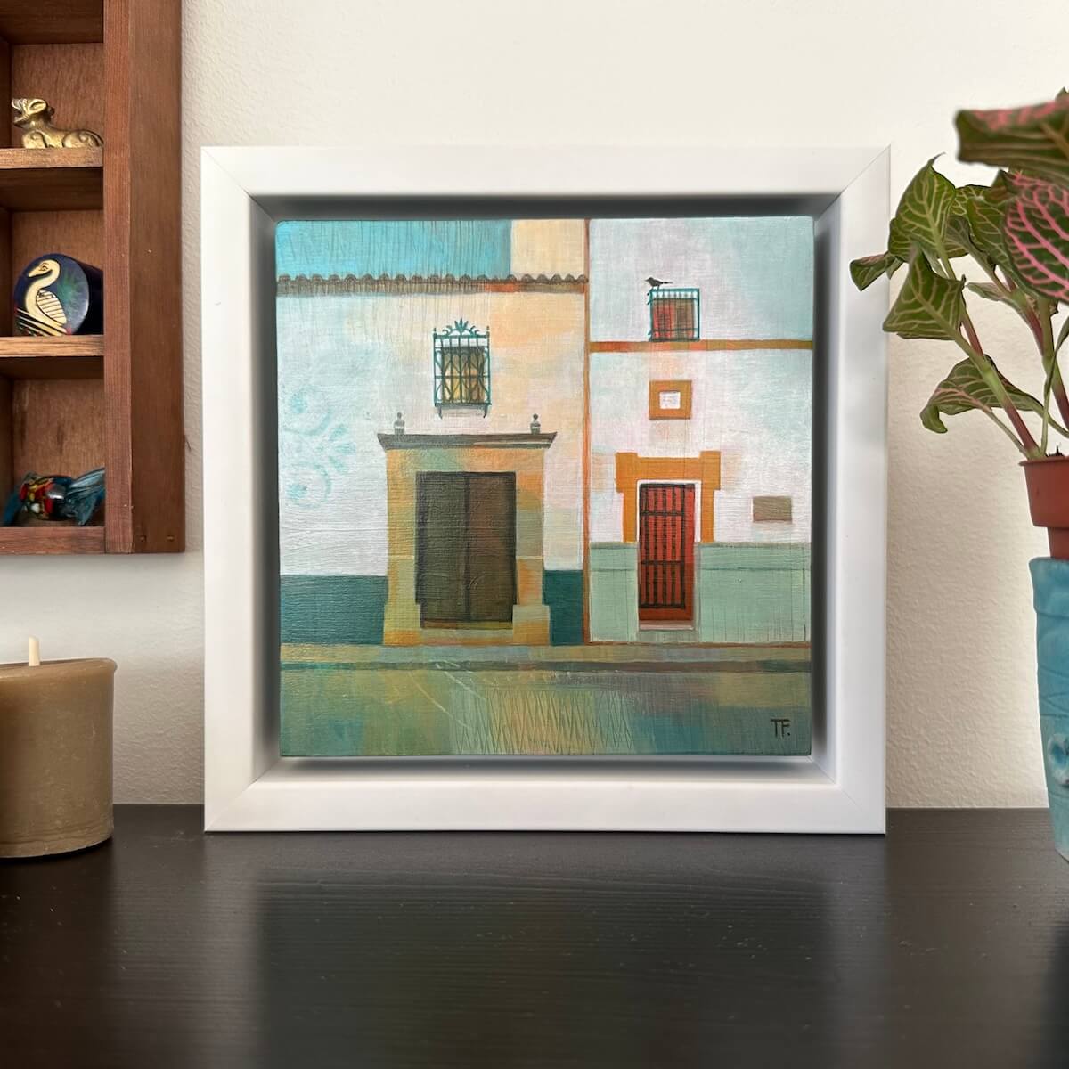 Framed artwork of a colourful Spanish street scene on a table with a plant and candle in the background.