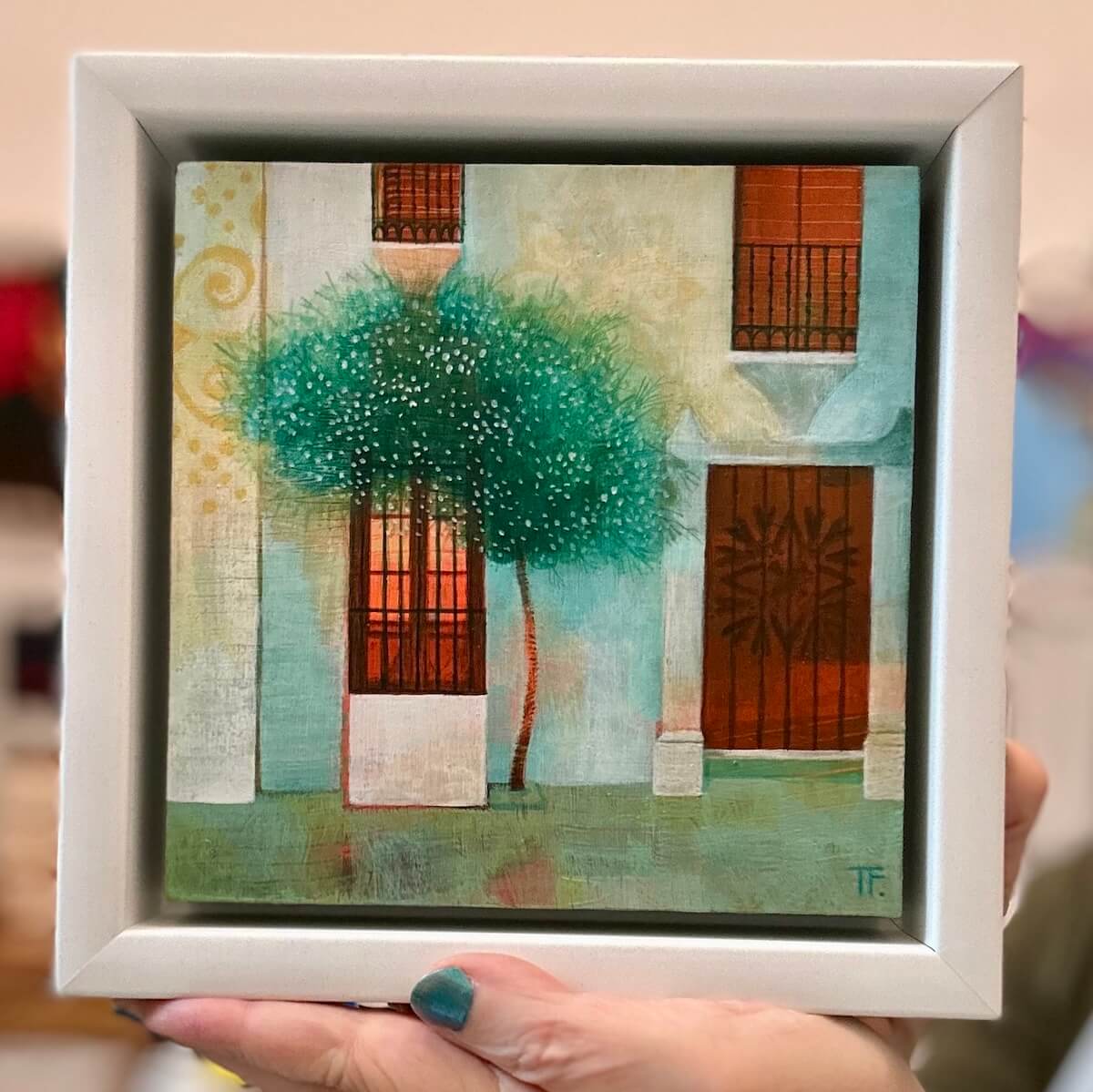 Framed artwork of a tree and Spanish building with red door held by a person.