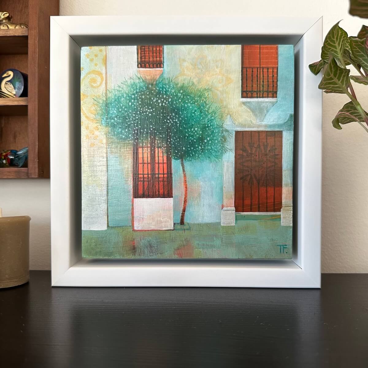 Framed abstract painting with Spanish house facade and a tree on a wooden surface.