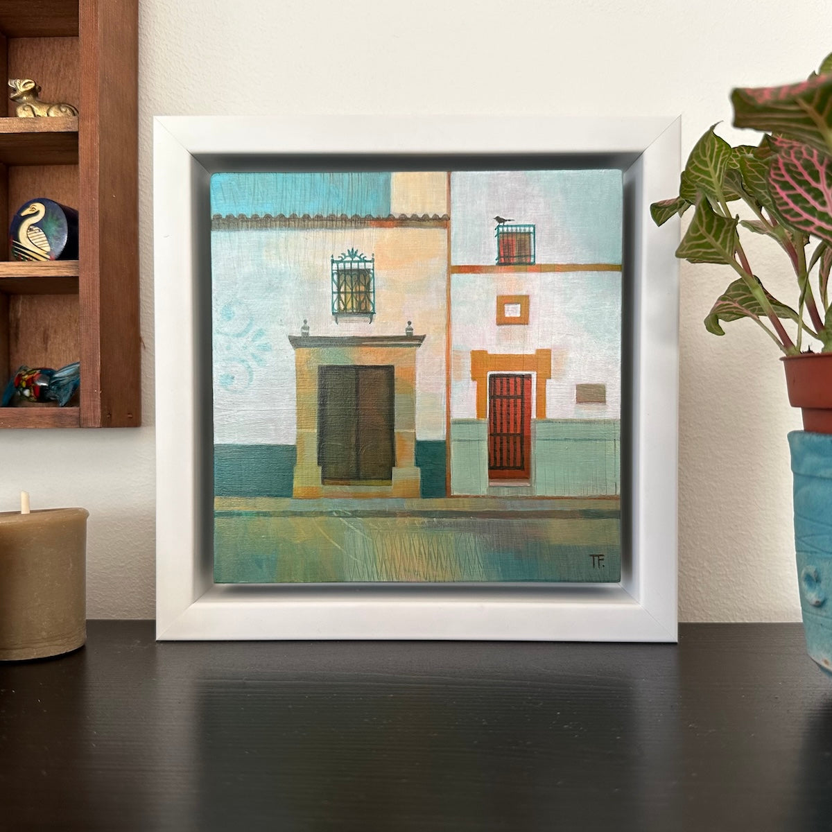 Framed artwork of a Spanish street scene on a wall with a plant and candle in the foreground.