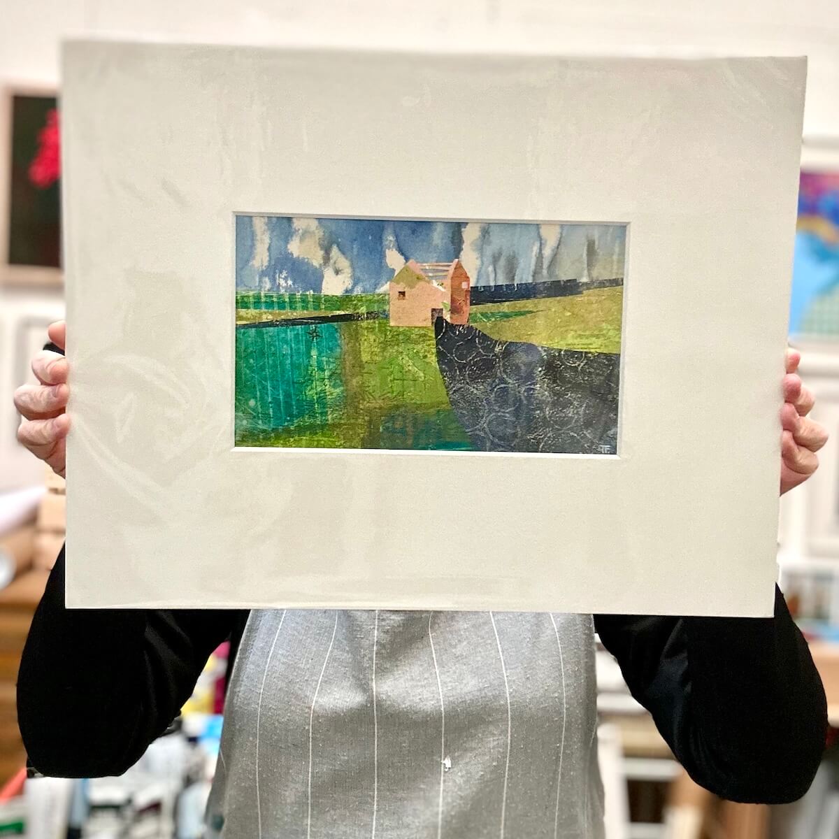 Person holding a mounted artwork of a landscape with a cottage.
